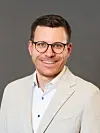 Portrett av smilende mann i lys jakke og briller foran nøytral bakgrunn.