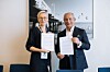 To personer innendørs holder signerte dokumenter foran et stort svart-hvitt-fotografi.